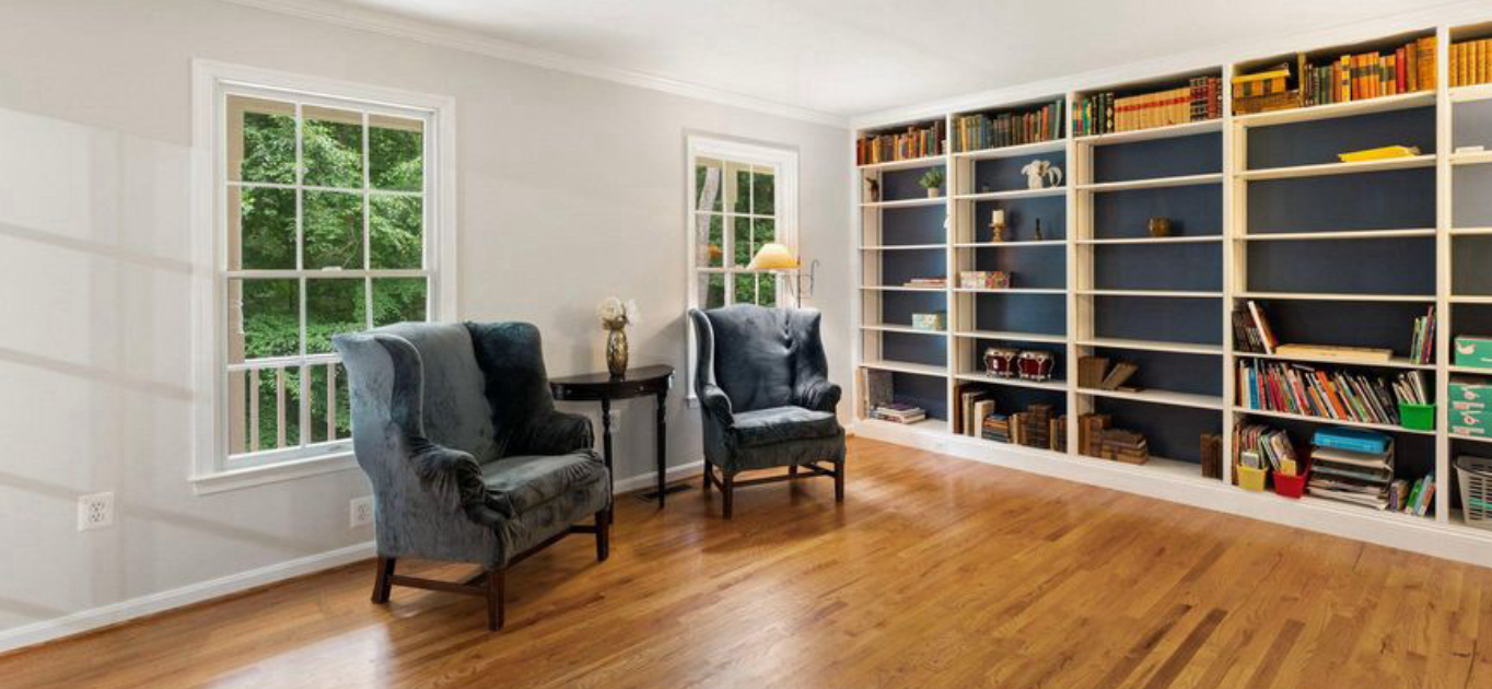 Well lit room with hardwood floors. Floor to ceiling built in bookshelves with white trim and blue back panel. Two blue wing chairs sit against the wall with windows behind. 